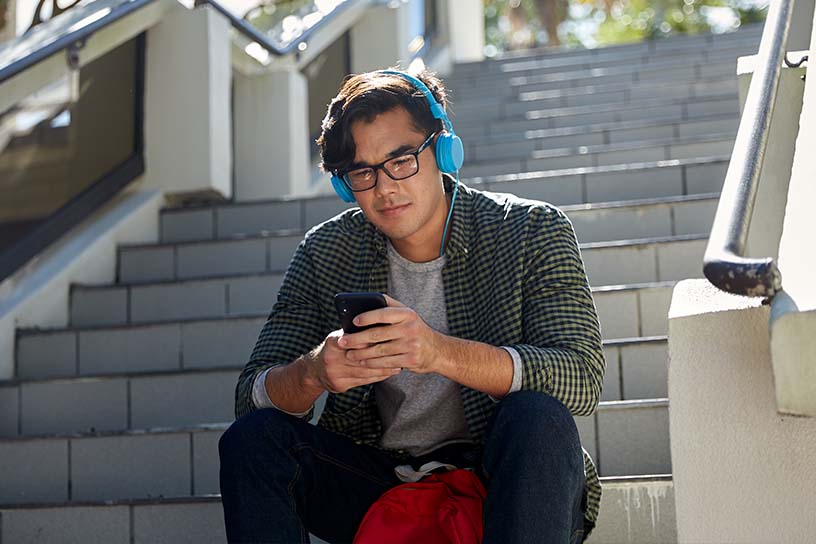 A young man sitting outside on a set of stairs, looking at this phone while wearing blue headphones. 