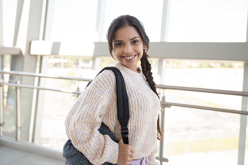 A young woman smiling toward the camera, wearing a backpack with one strap on her shoulder.