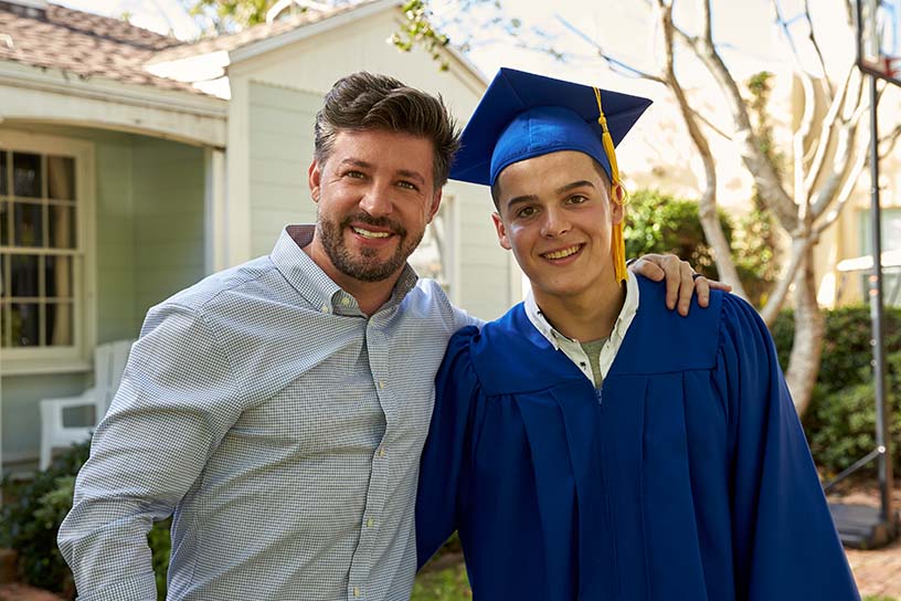 An older man standing next to a younger man who is wearing a blue graduation cap and gown; both have one arm wrapped around each other and are smiling toward the camera.